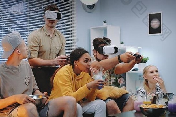 两个帅哥戴着虚拟现实眼镜，在欢快的同伴中玩游戏机游戏，他们一起吃零食，喝干冰鸡尾酒。