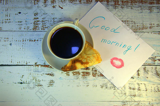 一杯咖啡，一个新烤的煎饼和一张纸，上面有一个早上好的愿望和一点口红。特写。