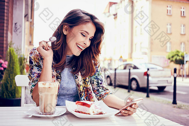快乐的年轻女子在户外咖啡馆吃蛋糕，并在智能手机上阅读信息