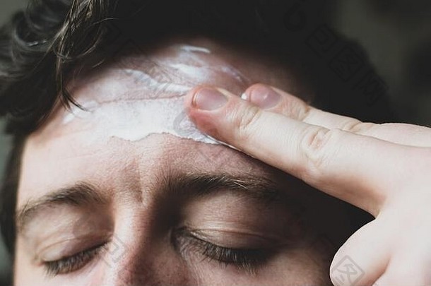 一个三十多岁的男人的脸部特写，作为日常护肤的一部分，他使用面霜滋润前额