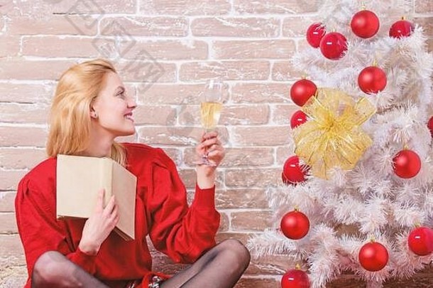 圣诞节庆祝概念。诺埃尔和乔伊。穿着红色连衣裙的女孩在圣诞树旁喝着香槟放松。女人高兴地在圣诞树旁喝酒，尽情庆祝。平安夜。