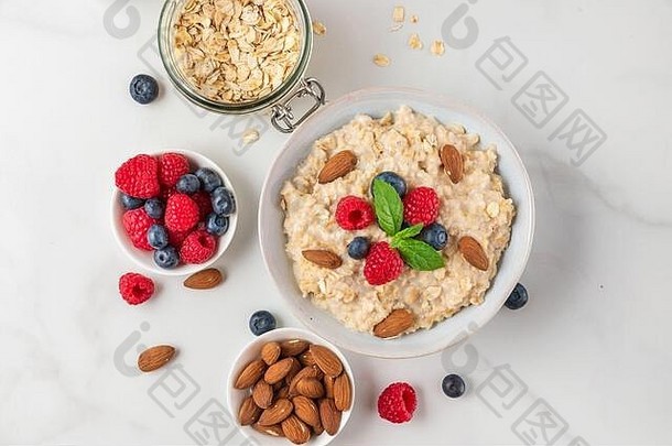 碗中的燕麦粥，白色背景上有新鲜浆果、坚果和薄荷，是健康饮食早餐。俯视图