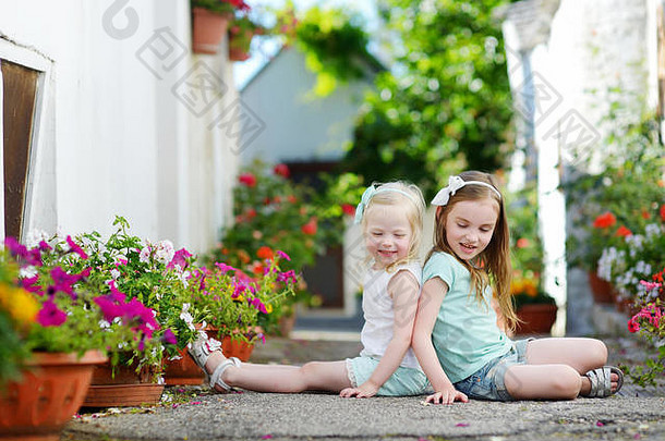 两个可爱的小姐妹坐在花盆之间，在温暖阳光明媚的夏日，在意大利小镇