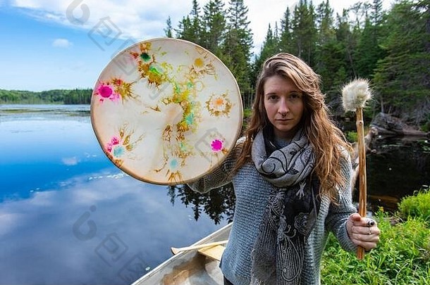 白天，在加拿大魁北克北部的湖滨，一位美丽的女子手持五颜六色的神圣的土制框架鼓和毛皮覆盖的鼓棒