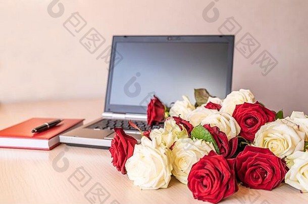 鲜花是工作场所新鲜白玫瑰和红玫瑰的礼物，背景略带粉红色。笔记本电脑键盘旁的木桌上放着大量的玫瑰。五、