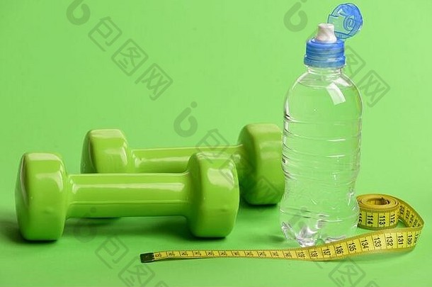 饮食和运动制度概念。瓶子旁边的塑料杠铃。亮绿色的哑铃，绿色背景上的水瓶和黄色卷尺。运动和保健设备