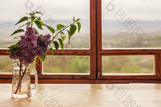 新鲜的紫丁香装在木窗前的玻璃瓶中，可以看到乡村景色。