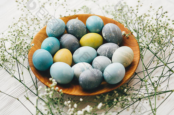 复活节快乐。白色木质背景上的木制盘子上有春天花朵的时尚复活节彩蛋。用天然染料涂成蓝色的现代复活节彩蛋