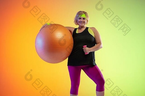 年轻的白人加大体型女模特在霓虹灯下的渐变橙色背景上训练。用健身球做运动。体育观念、健康生活方式、身体积极、平等。