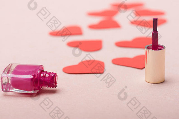 一瓶粉红色指甲油开着放在刷子旁边。许多纸心形的制作路径。浅粉色背景上的爱情象征，特写。情人节和美丽的概念。