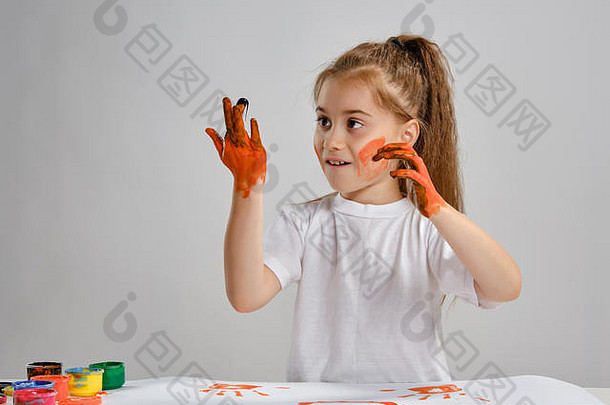 穿着白色t恤的小女孩坐在桌旁，桌上有惠特曼和五颜六色的颜料，在画她的手。白色的隔离带。中等特写镜头。