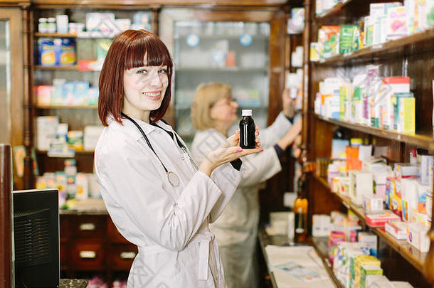 微笑迷人的年轻女士药剂师手里拿着一瓶药，专注于她的脸。另一名女药剂师在医院选择药物