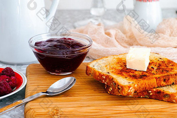 两杯烤面包，木板上放一块黄油，桌上放一碗覆盆子酱。特写。
