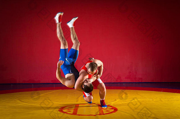 两个穿着蓝色和红色摔跤紧身衣的年轻人正在体育馆的黄色摔跤地毯上进行摔跤和超级摔跤