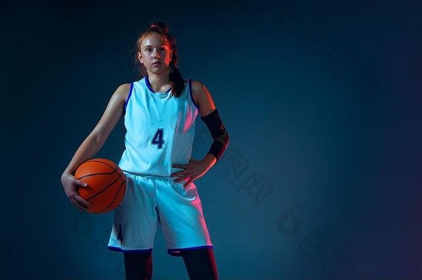 强烈年轻的白人女篮球运动员，在霓虹灯、动作和动作的蓝色工作室背景下。运动、运动、活力和健康生活方式的概念。摆出自信的姿势。