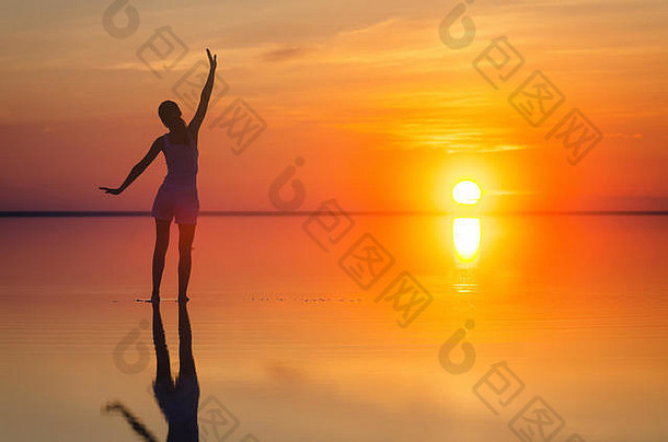 美丽的女模特在海边的日出下张开双臂。盐湖埃尔顿平静的海水折射出女人的轮廓。太阳落在地平线后面。那个女孩独自一人