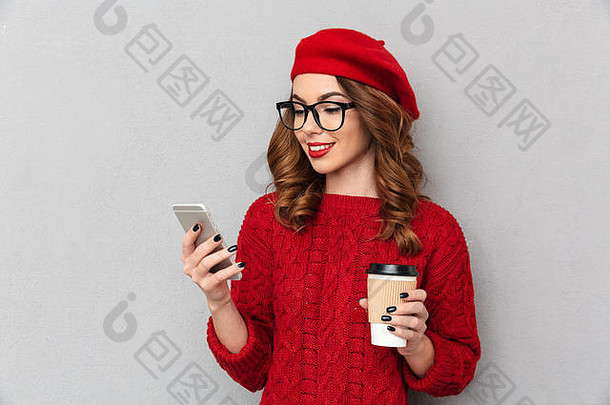 一位穿着红色毛衣、戴着眼镜、手持手机、隔着灰色墙壁背景拿着外卖咖啡杯的快乐女人的肖像