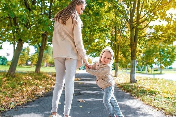 女人妈妈走路，牵着小男孩的手，秋天在城市里，漫步在公园里，草地背景，温暖的休闲服，带风帽的米色开衫