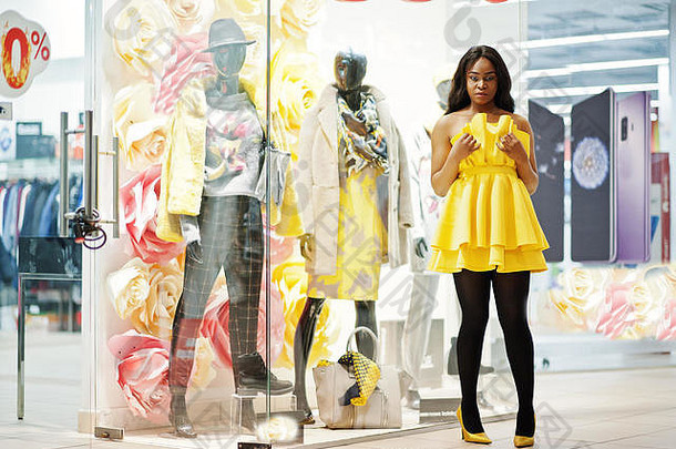 穿着黄色睡衣的时尚非洲裔<strong>美</strong>国妇女在<strong>商场</strong>里与模特儿对着<strong>陈</strong>列柜摆姿势。