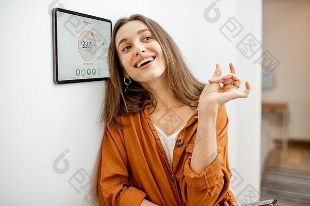 一位年轻快乐的女士的肖像，靠近智能<strong>家居</strong>控制的触摸屏面板。简单方便的家庭智能设备管理应用程序的概念