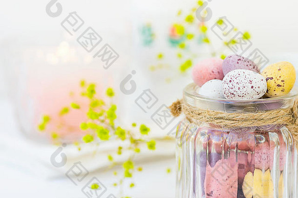 巧克力糖果多色小鹌鹑复活节彩蛋彩色复古玻璃罐用绳系在白色木桌上黄色春花点亮