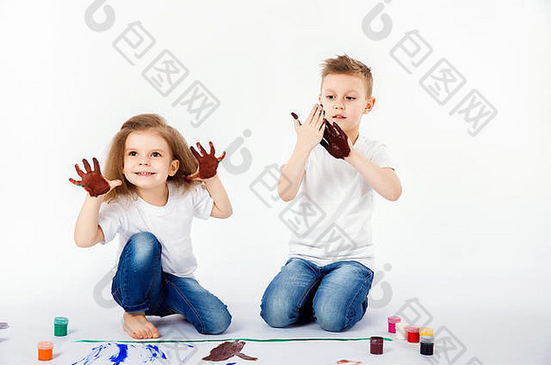 两个穿着白衬衫和蓝色牛仔裤的可爱的儿童朋友，一个男孩和一个女孩，时髦的发型，赤脚，用白色的颜料在白色的纸上<strong>画画</strong>。在油漆上举手。