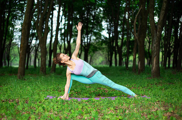 苗条的黑发女孩在一个夏季公园里做运动，做美丽而复杂的瑜伽姿势。背景是郁郁葱葱的绿色森林。女人在做什么