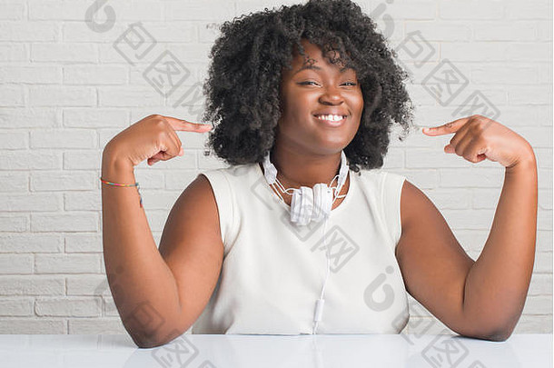一位年轻的非洲裔美国妇女戴着耳机坐在桌子上，脸上带着自信的微笑，用手指指着自己，骄傲而快乐