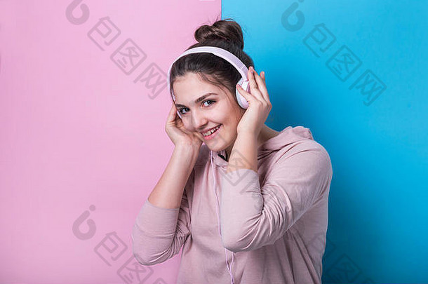 快乐的少女戴着粉色耳机，在彩色画室背景前微笑着听音乐。青年与青年文化
