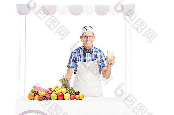 一个老苏打混蛋，手里拿着一杯柠檬水，站在一个摊位后面，摊位上放着一堆水果