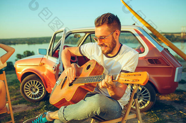 这对情侣在河边的一个夏日里坐在沙滩上弹吉他休息
