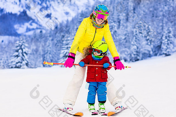 母亲和小孩在阿尔卑斯山滑雪。带安全帽、护目镜和杆子的活跃的妈妈和蹒跚学步的孩子。滑雪课