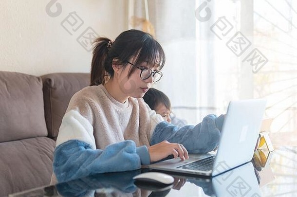 在冠状病毒大流行期间，一位年轻的亚洲母亲在家里用现代笔记本电脑工作，她的孩子在平板电脑上看动画片