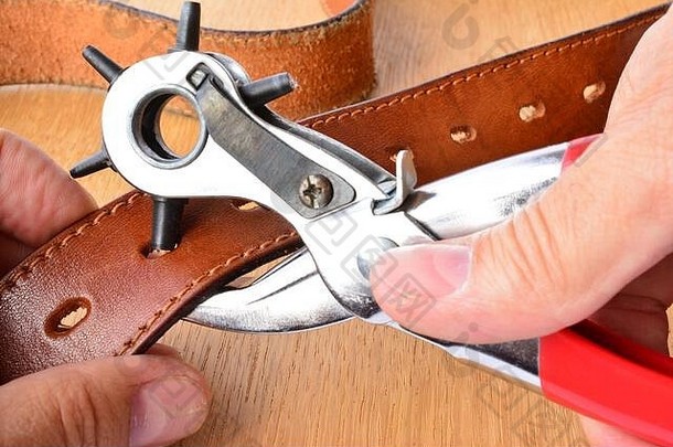 皮革腰带穿孔，冲压工具和新的皮革腰带在工匠的手在木制背景