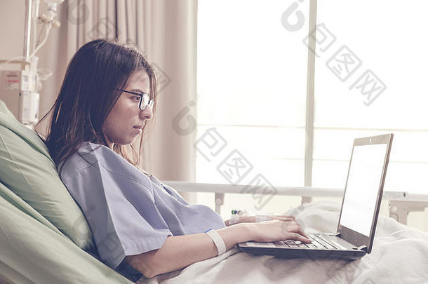 年轻的亚洲女病人躺在病床上，在医院康复期间使用笔记本电脑工作。healthc