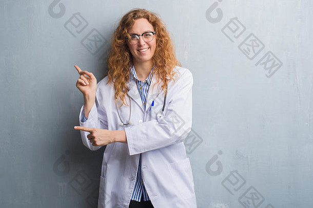 年轻的红发女医生站在灰色的垃圾墙上，身穿外套，微笑着看着摄像机，双手和手指指向侧面。