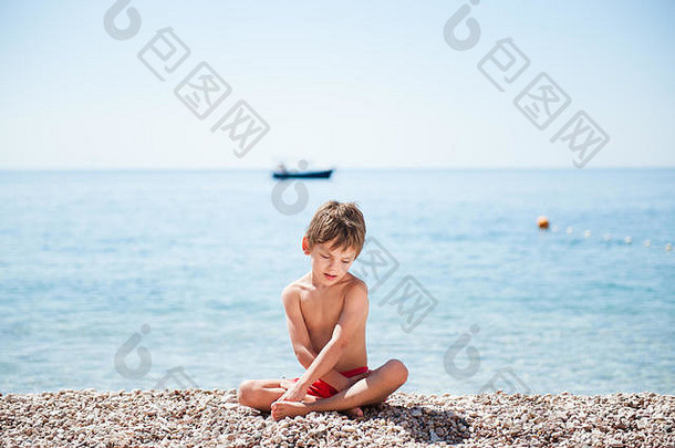英俊的白人小男孩坐在海边的海滩上，身后有一条小船