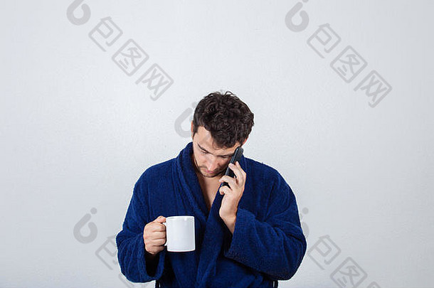 一幅困倦的年轻商人的画像，穿着蓝色浴袍，从杯子里喝咖啡，在白色背景下对着电话讲话。