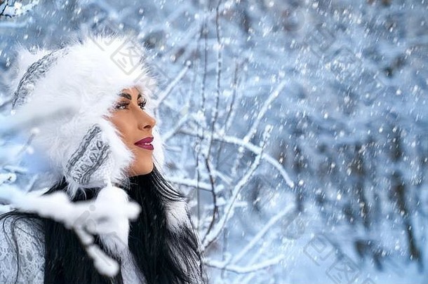 戴着暖冬帽的美丽女孩在覆盖着雪的树旁的冬日里摆姿势。正模远眺，欣赏冬日阳光明媚的大自然。<strong>冬季</strong>服装的概念。