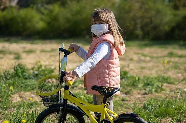 阳光明媚的日子里，一个戴着口罩的小女孩骑自行车。孩子们，在疫情期间玩耍。儿童冠状病毒防护。