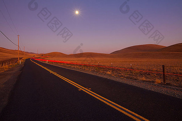 黄昏时浪漫的乡间小路。一辆过往汽车的尾灯消失在远处。蓝色，紫色的天空，升起的月亮。
