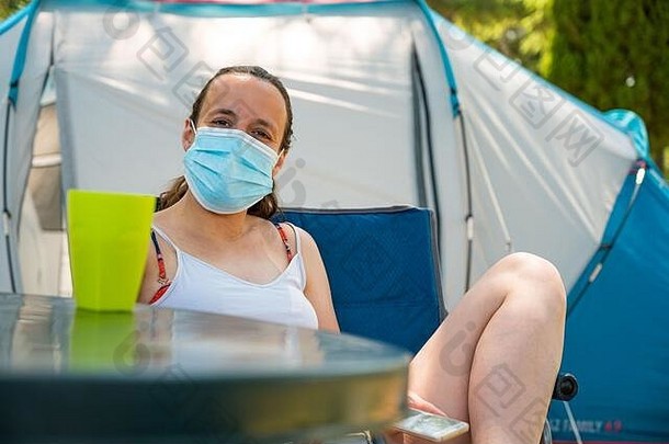 在一个度假胜地，一名戴着医用面罩的妇女坐在露营帐篷前。