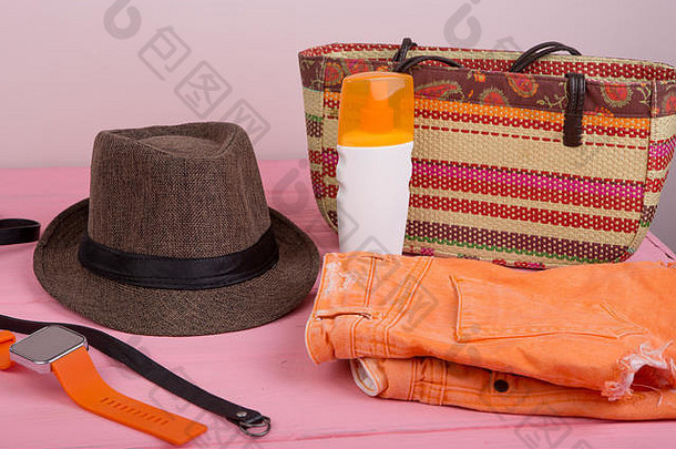 夏季配饰-稻草沙滩包、太阳帽、腰带、橙色手表、牛仔短裤、粉色木桌上的防晒霜