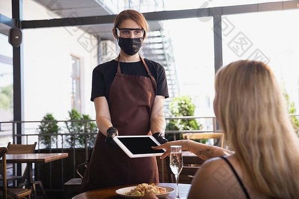 在冠状病毒大流行期间，这位女服务员戴着医用口罩、手套在餐厅工作。代表服务和<strong>安全</strong>的新常态。非接触式支付，照顾客户。