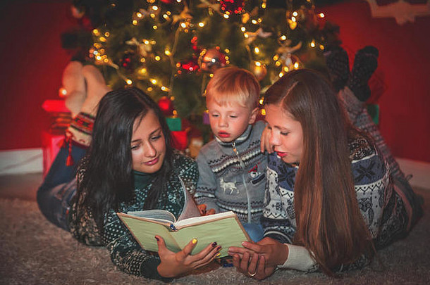 年轻的家庭和孩子在装饰过的房间里的圣诞树旁读这本书。
