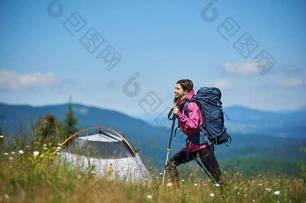 运动型女登山者，背着蓝色背包，在帐篷附近拄着登山杖，穿着运动服，在蓝天和群山映衬下的山顶上徒步旅行，微笑着，享受着夏日的早晨
