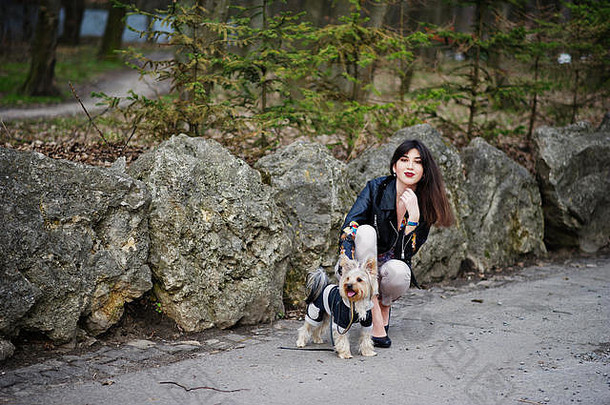 深色头发的吉普赛女孩和约克郡猎犬在公园的石头上摆姿势。模特穿皮夹克和t恤，带饰物，裤子和鞋子w