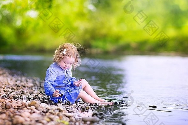 可爱的卷曲的蹒跚学步的女孩穿蓝色的衣服玩河海岸<strong>扔石头</strong>水热夏天一天