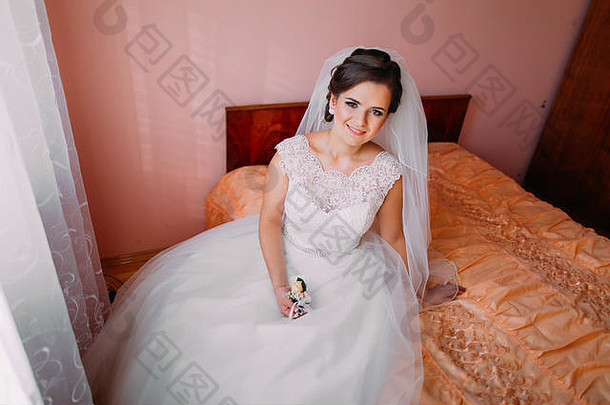 美丽的新娘坐在靠近窗户的床上等待，手里拿着可爱的<strong>小结</strong>婚花束