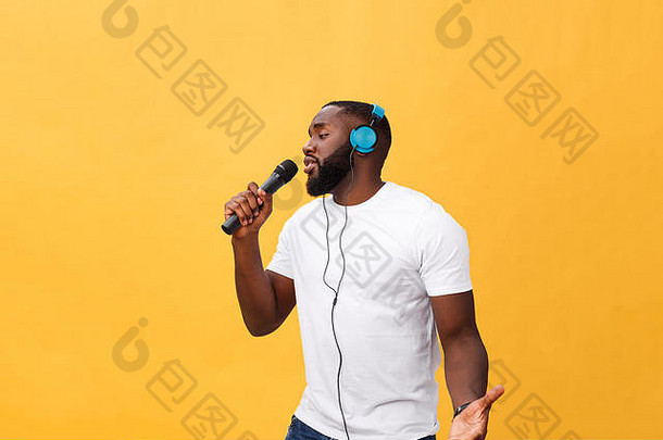 照片中是一位快乐、积极、时尚、英俊的非洲男子，他手持麦克风，头戴耳机，在黄色背景下听着音乐唱歌，享受周末假期